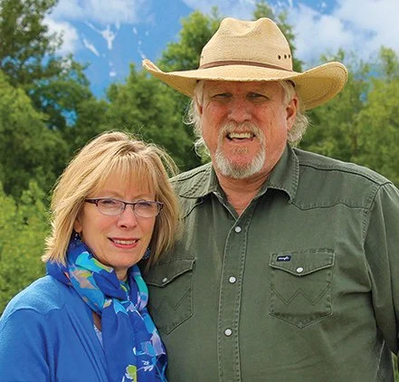 A smiling woman wearing glasses, a blue jacket, and a blue scarf, standing next to a smiling man in a green shirt and a cowboy hat, outdoors with green trees and blue sky in the background.