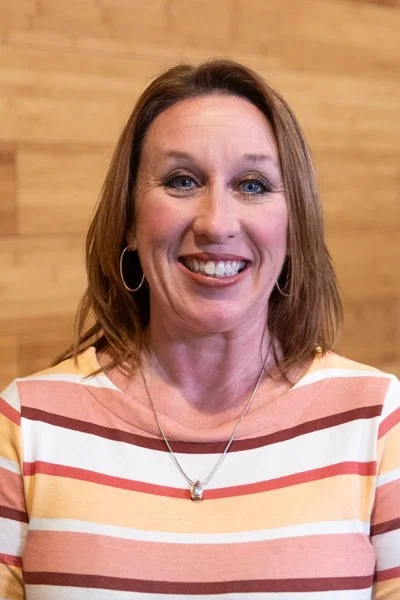 A woman smiling with shoulder-length brown hair, wearing a striped shirt and a necklace, standing in front of a wooden background.