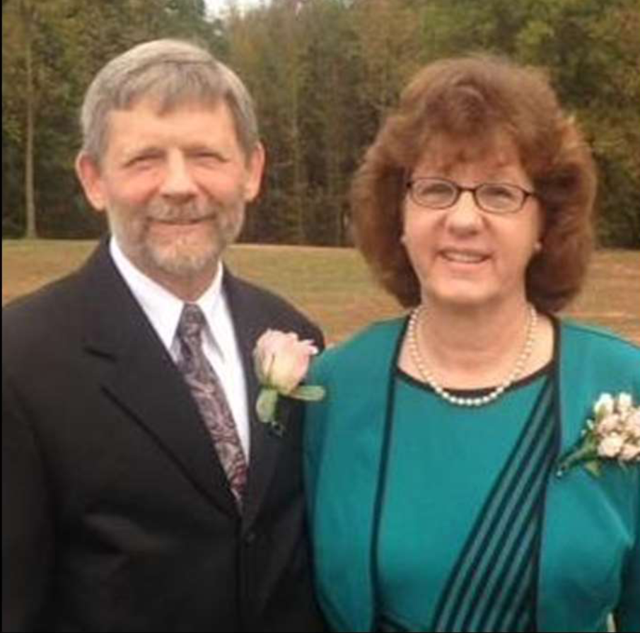 A middle-aged man with a beard and a middle-aged woman with glasses, both dressed formally, are standing outdoors on a grassy field with trees in the background. They are smiling and wearing corsages.