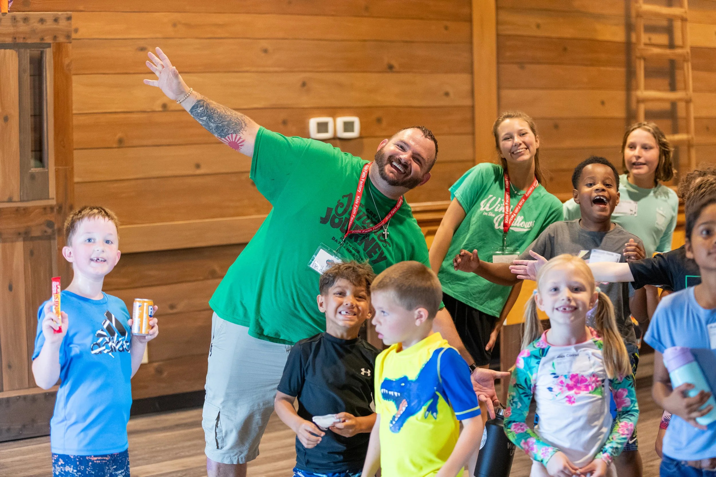 A group of children and two adults smiling and having fun indoors with wooden walls in the background.