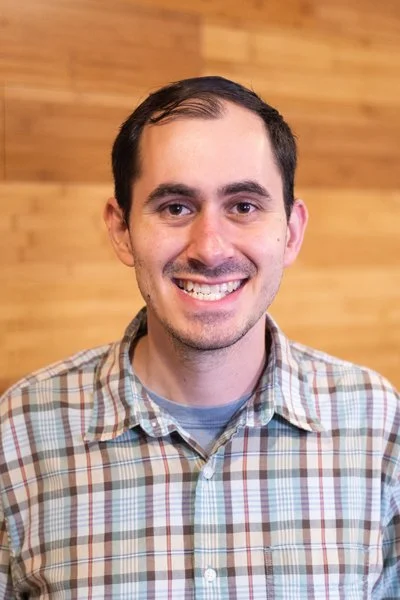 Smiling man with short dark hair, wearing a plaid shirt, standing in front of a wooden wall background.