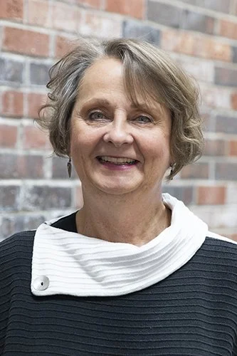A smiling older woman with short, wavy gray hair, wearing a black top and a white scarf, standing in front of a brick wall.