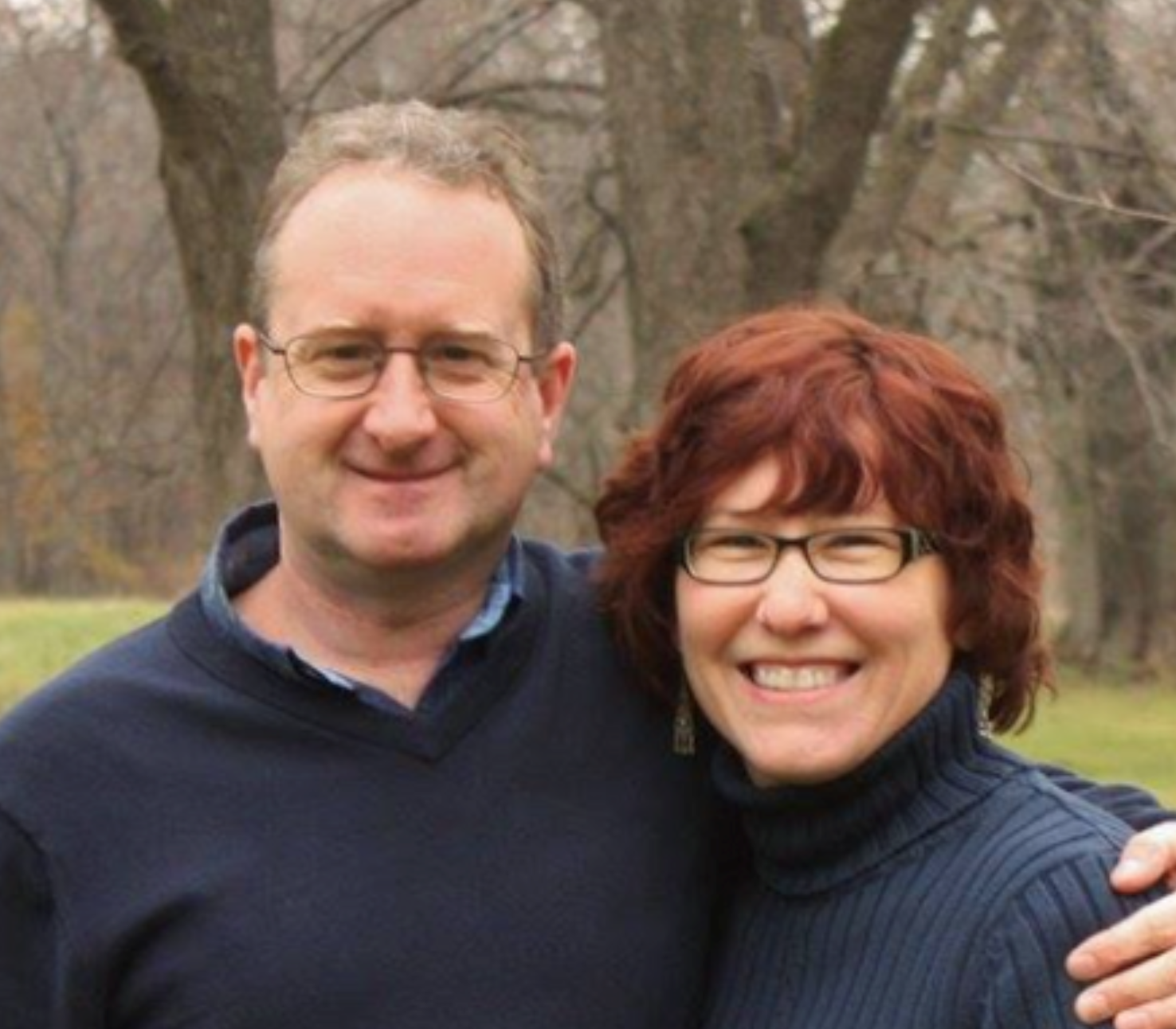 A smiling man and woman with glasses standing outdoors in front of leafless trees on a cloudy day.