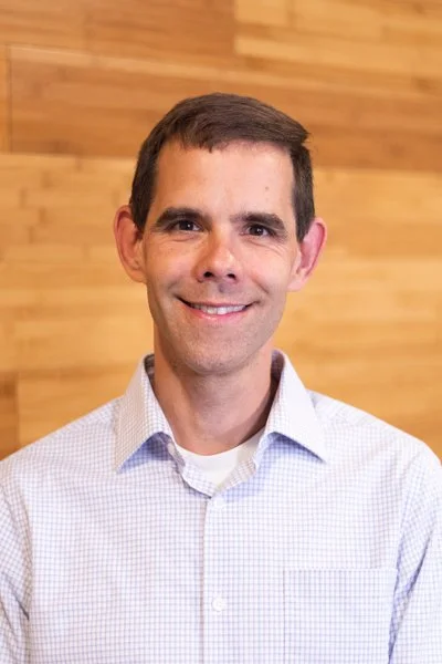 Portrait of a man smiling, wearing a checkered shirt, with a wooden wall background.