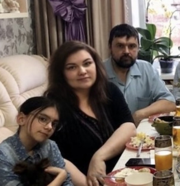 A family of three sitting at a dining table, with plates of food and drinks in front of them. The woman in the middle has long dark hair, and the man on her right has a beard and glasses. The girl on her left wears glasses and has long dark hair.