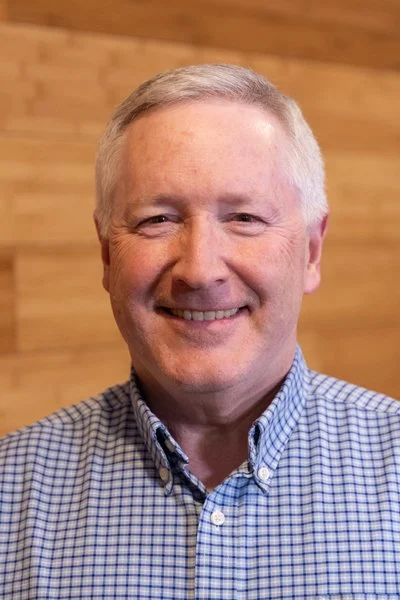 A middle-aged man with short gray hair and a broad smile, wearing a blue checkered button-up shirt, standing in front of a wooden background.