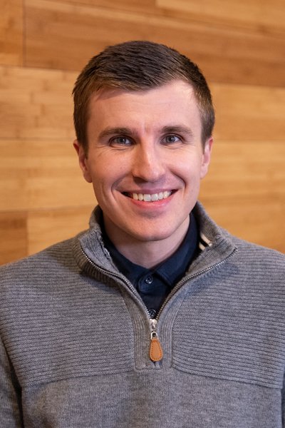A smiling man with short brown hair, wearing a gray zip-up sweater over a dark collared shirt, standing in front of a wooden wall background.