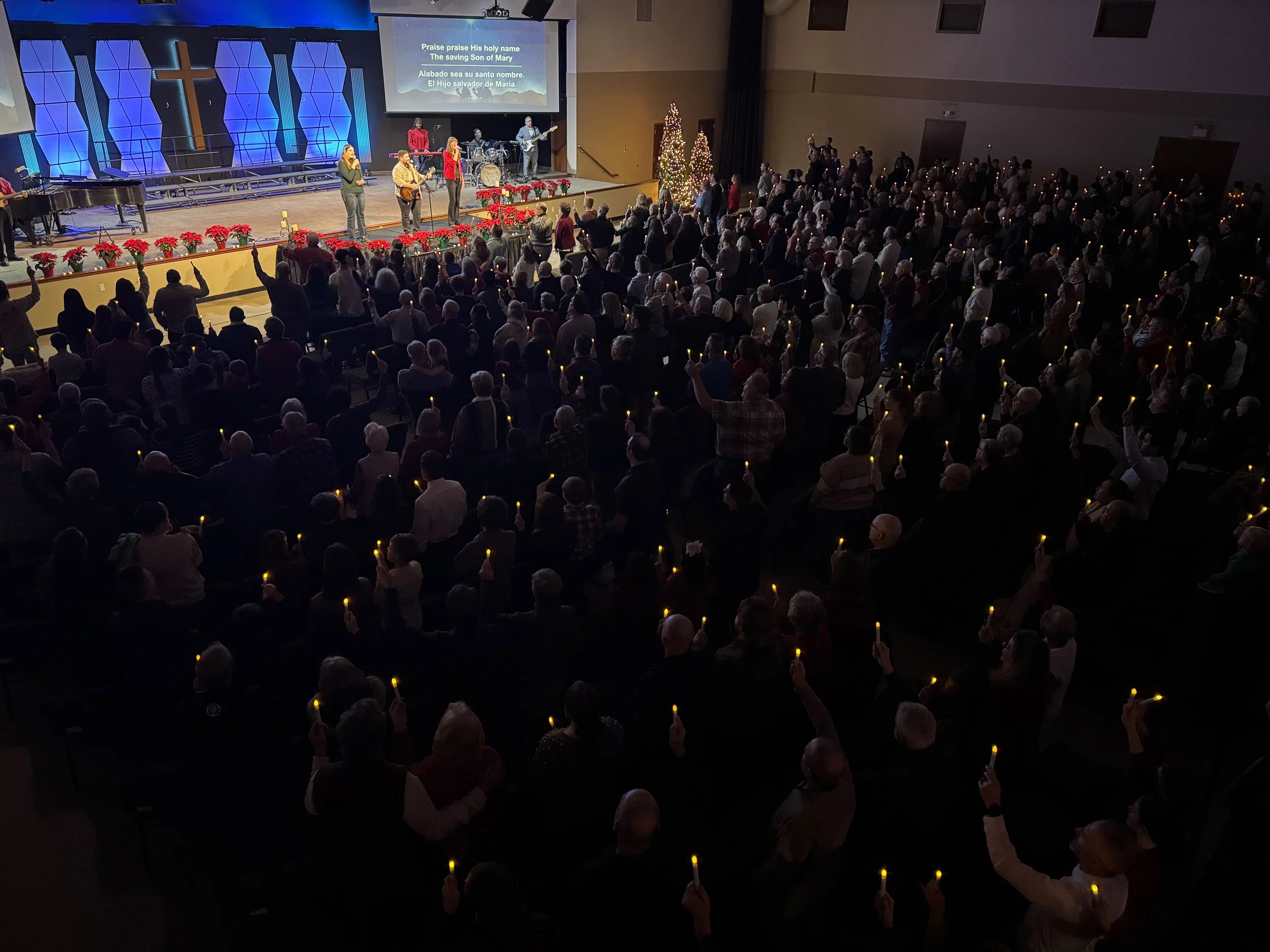 A large indoor church worship service with many attendees holding lit candles, facing a stage where a band and choir are performing. The stage is decorated with poinsettias and Christmas trees, and a screen displays lyrics in English and Spanish.