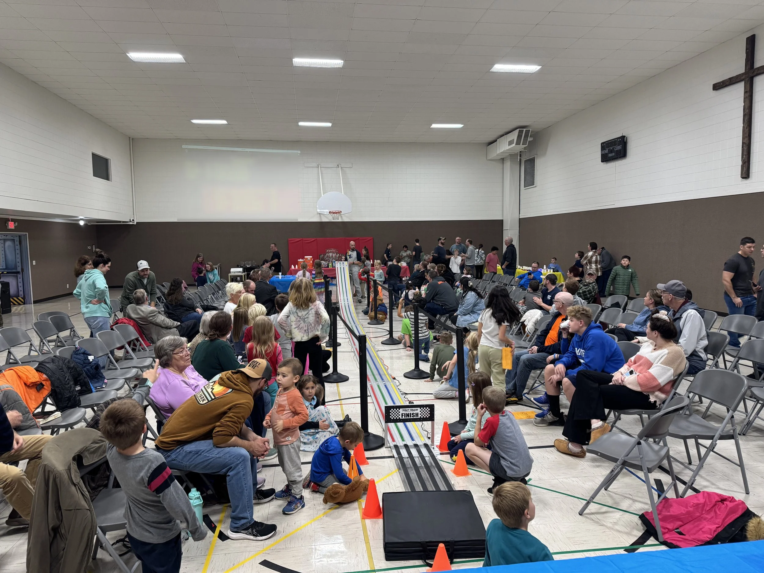 Children and adults gathered around a colorful miniature race track set up in a gymnasium for a race event, with some seated and others standing or walking.