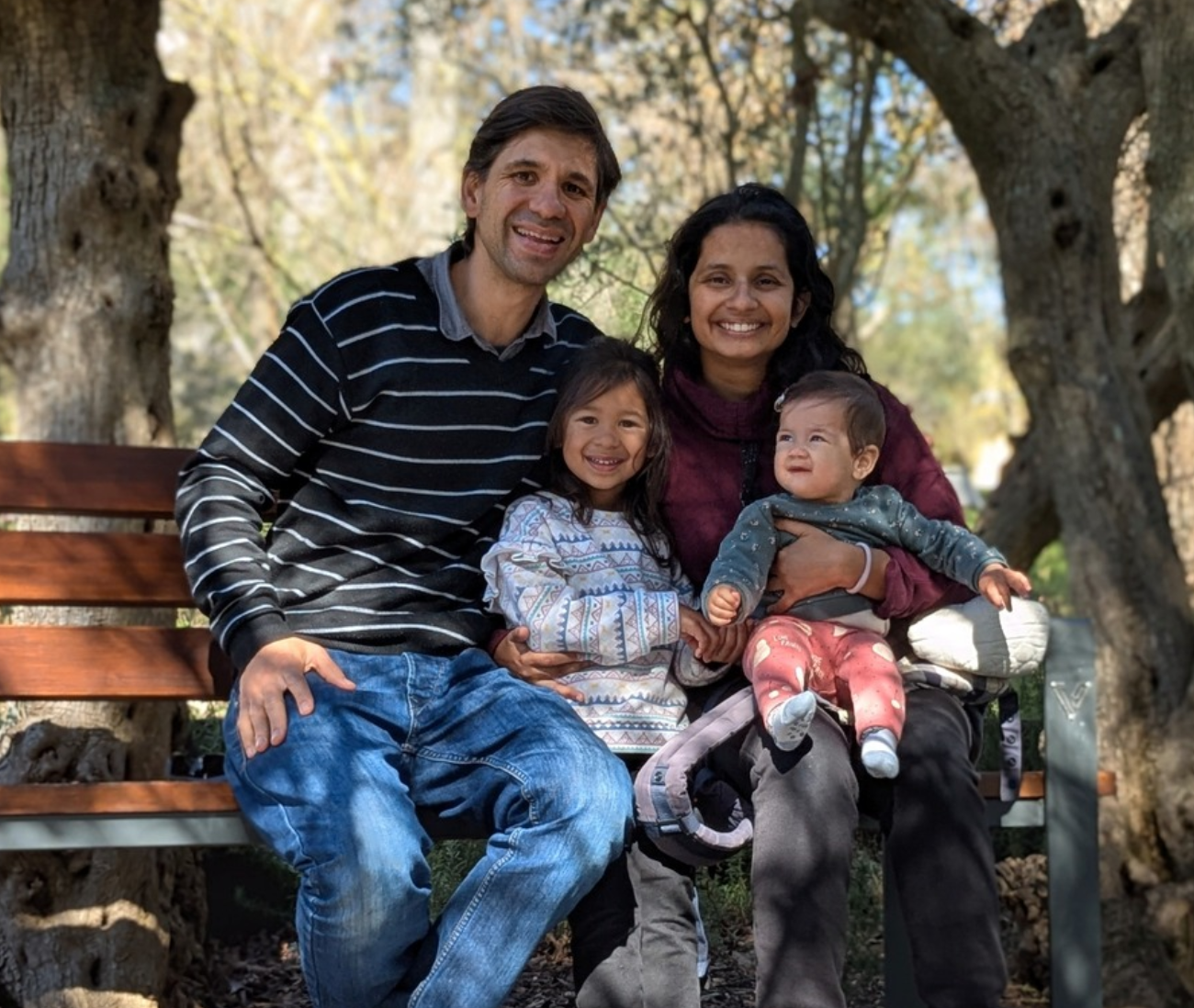 A family of four sitting on a park bench, smiling, with trees in the background.