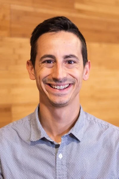A man smiling, with short dark hair, wearing a light gray collared shirt, in front of a wooden wall background.