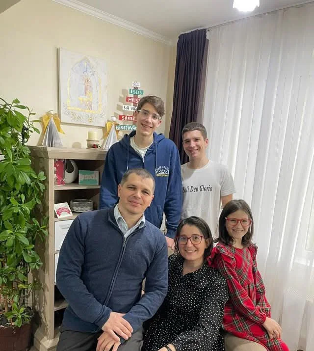 Five people, two women and three men, posing for a photo in a living room. The background has a bookshelf, a plant, and a curtain. Four are sitting, one is standing behind them.
