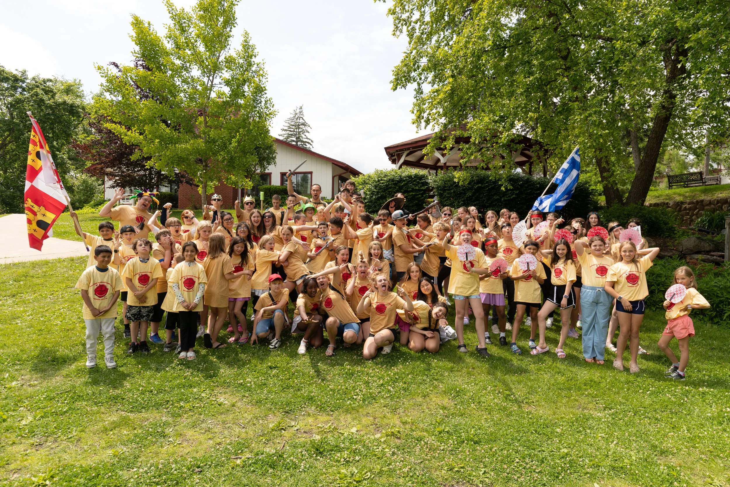 A large group of children and adults gathered outdoors on a grassy area, wearing matching yellow T-shirts, some holding fans and flags, with trees and houses in the background.