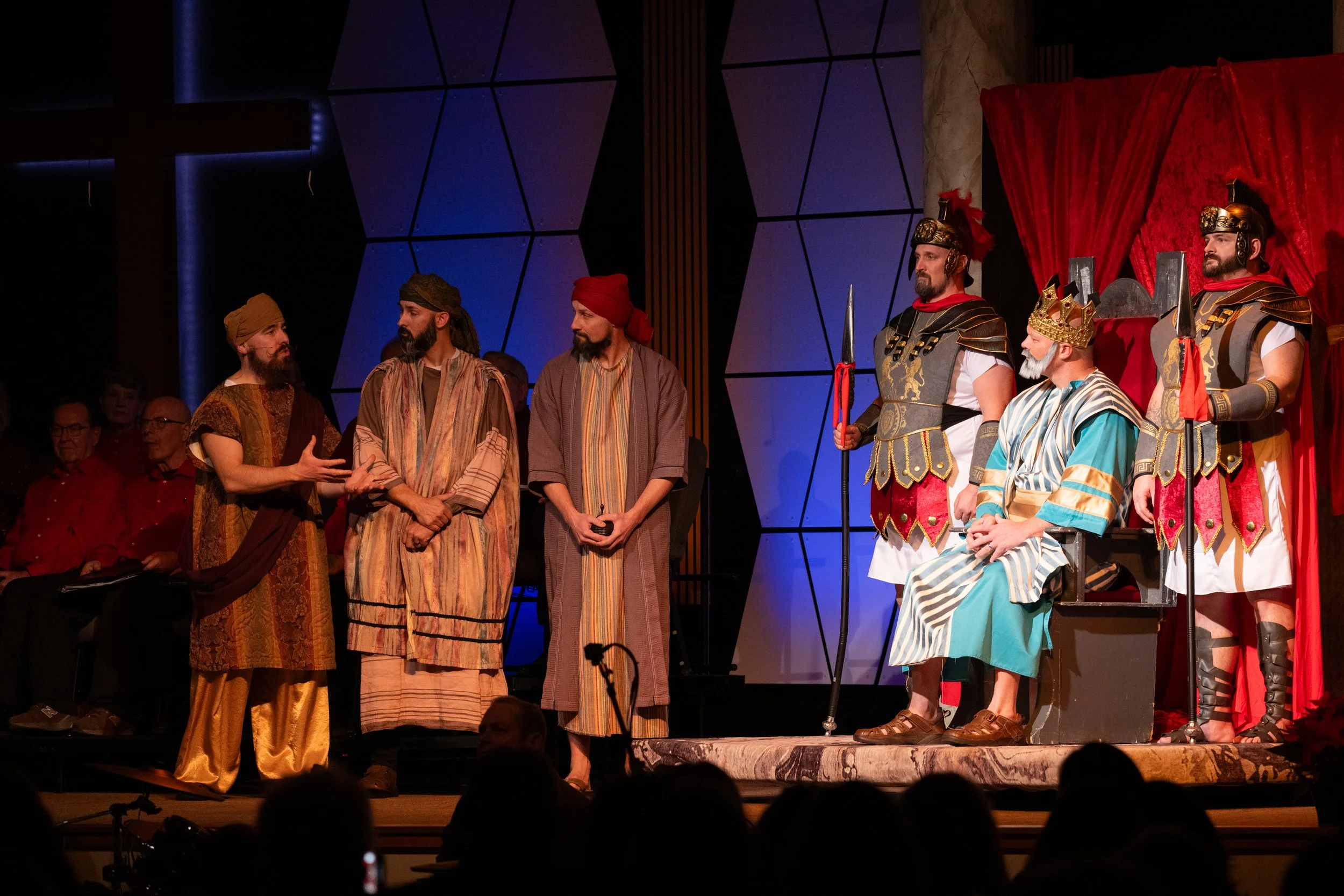 Actors in medieval costumes performing on stage with a red curtain and a geometric patterned backdrop.