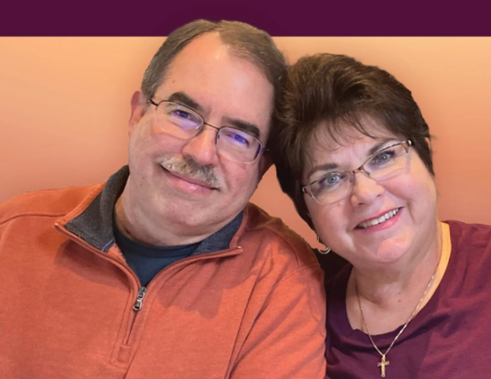 A middle-aged man and woman smiling and taking a selfie together, both wearing glasses and casual clothing, sitting against a peach-colored background.