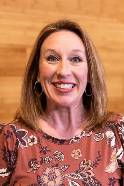 A woman smiling with shoulder-length brown hair, wearing a striped shirt and a necklace, standing in front of a wooden background.