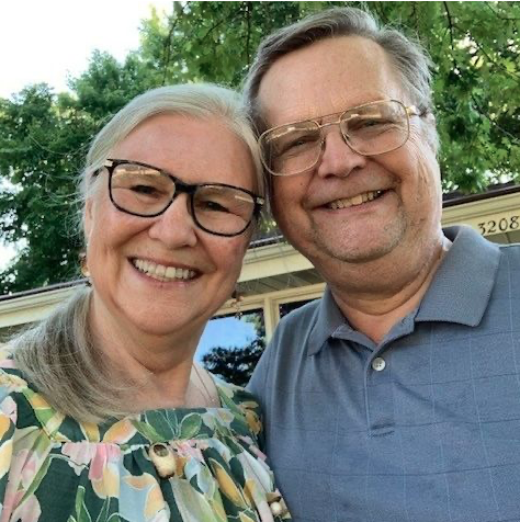 Smiling older man and woman taking a selfie outdoors with trees and a bus in the background.