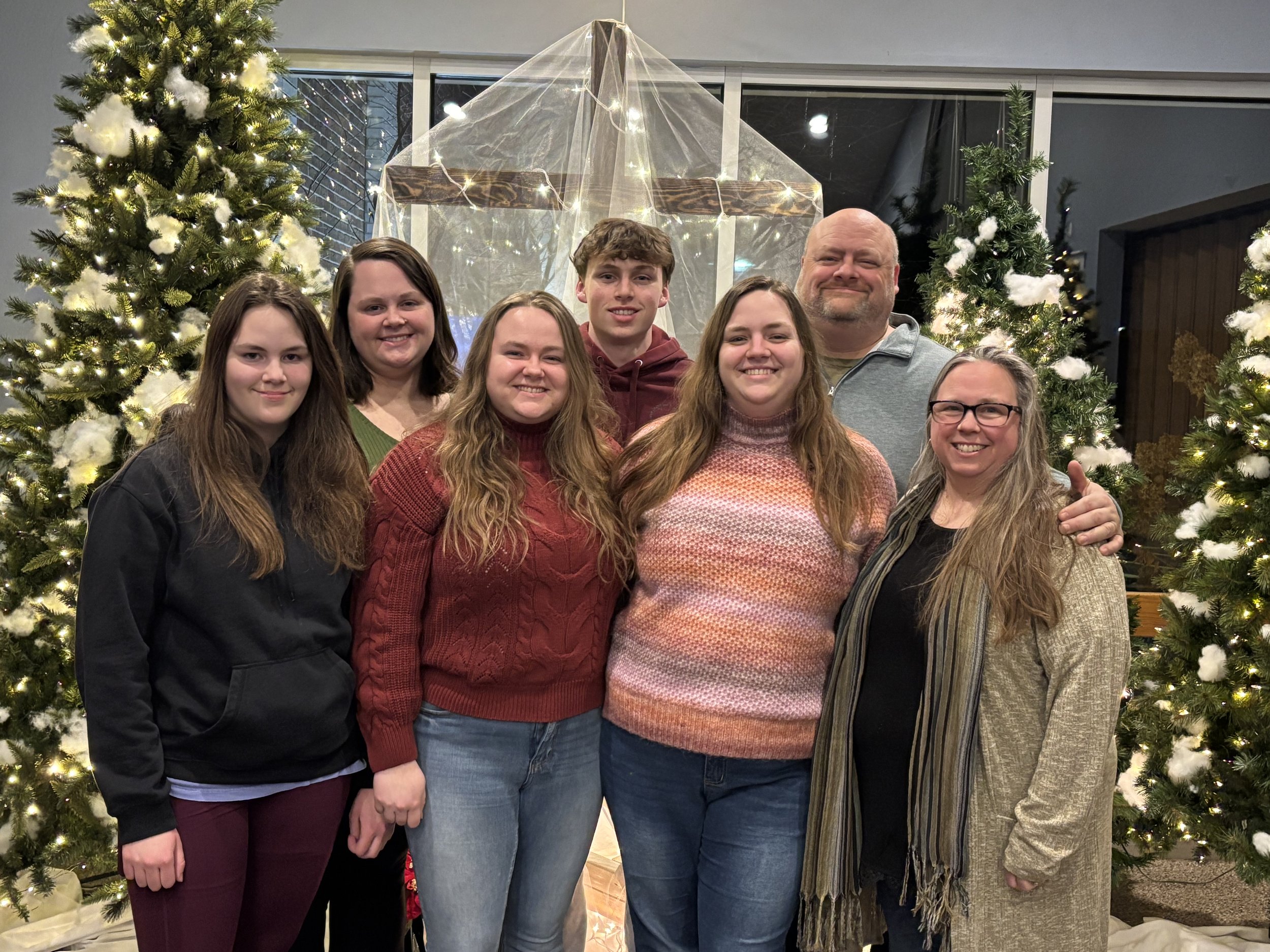 Group of seven people, including five women and two men, smiling and standing close together in front of decorated Christmas trees and a nativity scene with a clear covering and lights.