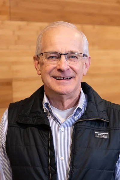 A smiling older man with glasses, wearing a striped button-up shirt and a black vest, standing in front of a wooden wall.
