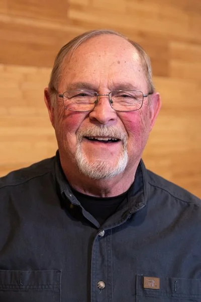 A smiling elderly man with glasses, a beard, and a mustache, wearing a black shirt, standing in front of a wooden wall.