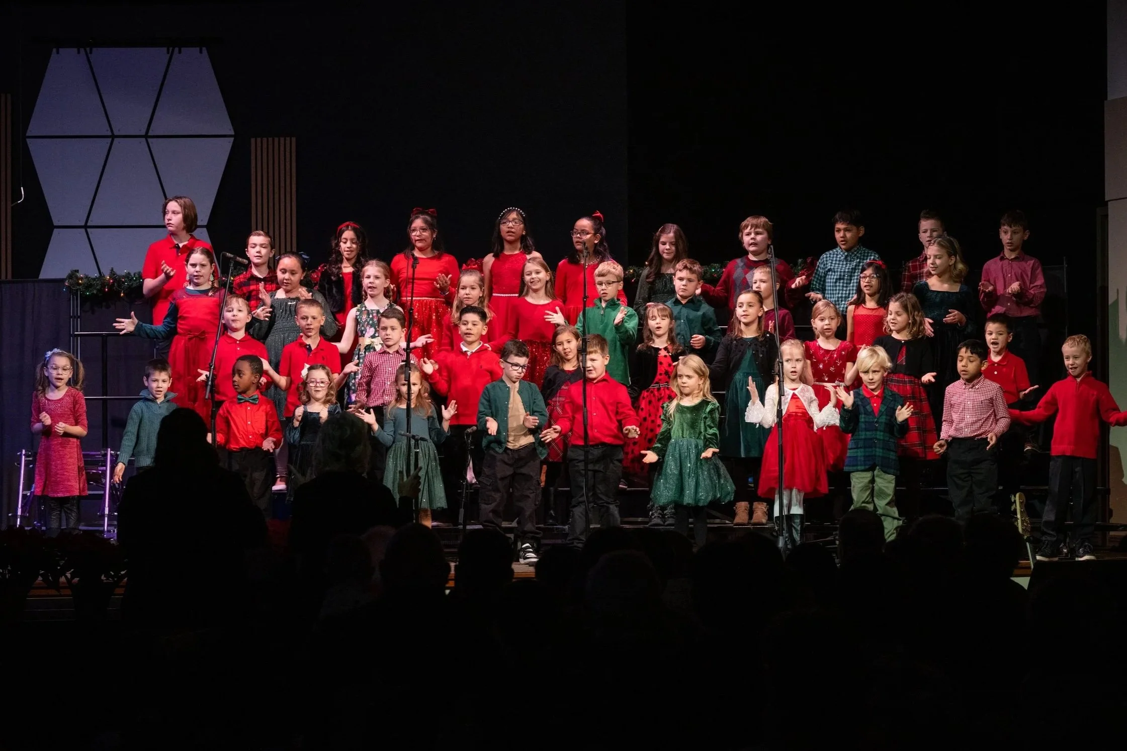 Children dressed in festive holiday attire singing on stage during a Christmas concert.