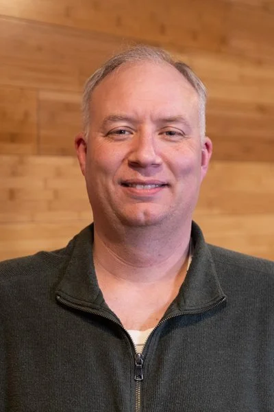 A man smiling, wearing a dark zip-up sweater, standing in front of a wooden wall.