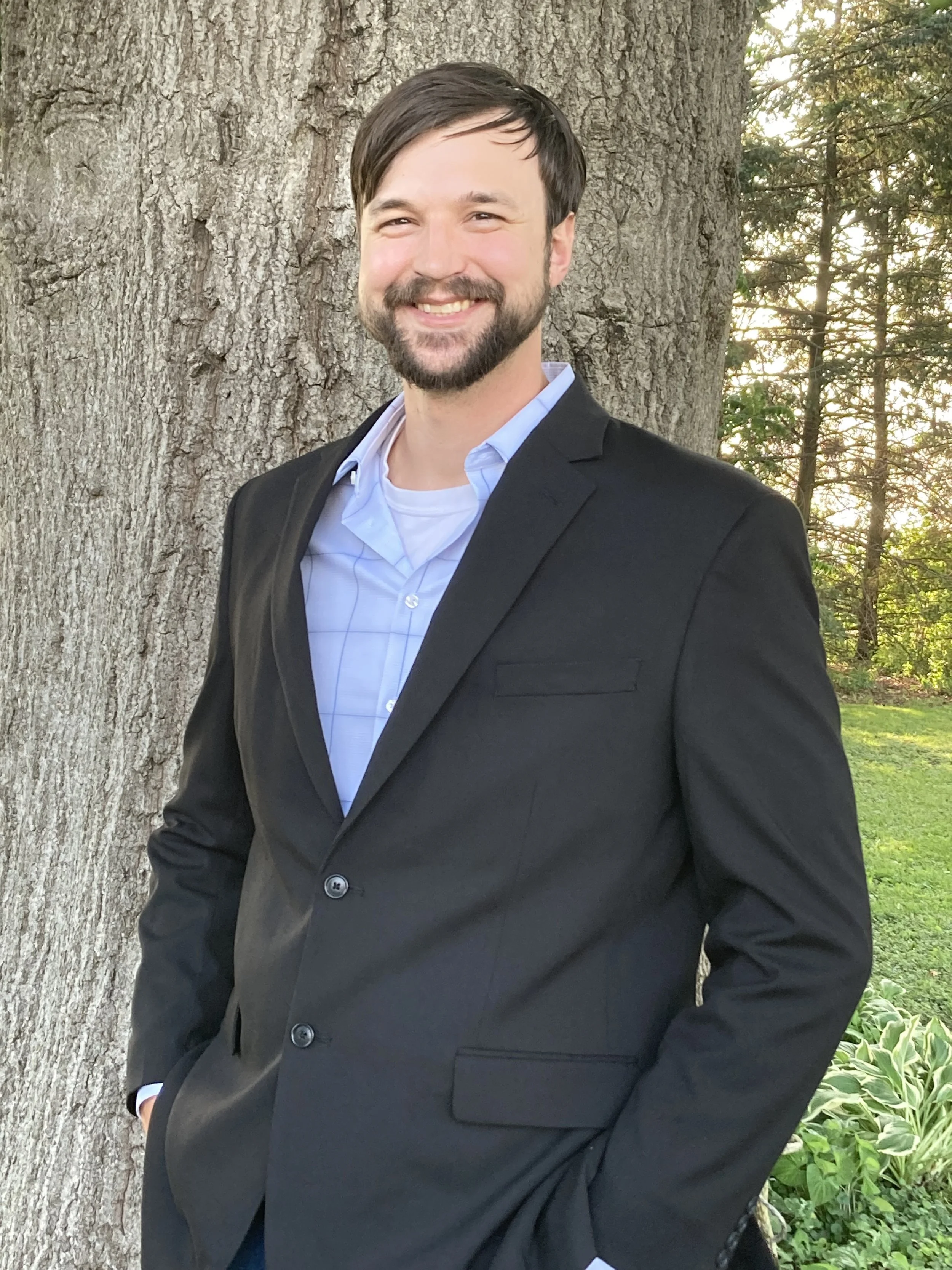 A man with dark hair and a beard, dressed in a dark suit and light blue shirt, is smiling and standing outdoors next to a large tree, with sunlight filtering through the trees in the background.