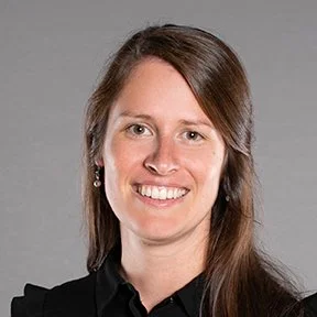 Portrait of a woman with long brown hair, smiling, wearing a black top and earrings, neutral gray background.