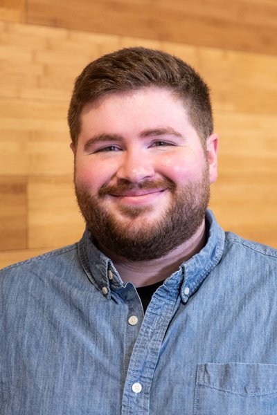 A young man with a beard and short hair, smiling, wearing a denim shirt, standing in front of a wooden wall.