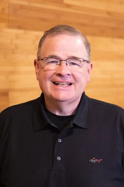 A smiling man with glasses wearing a black collared shirt, standing in front of a wooden wall.