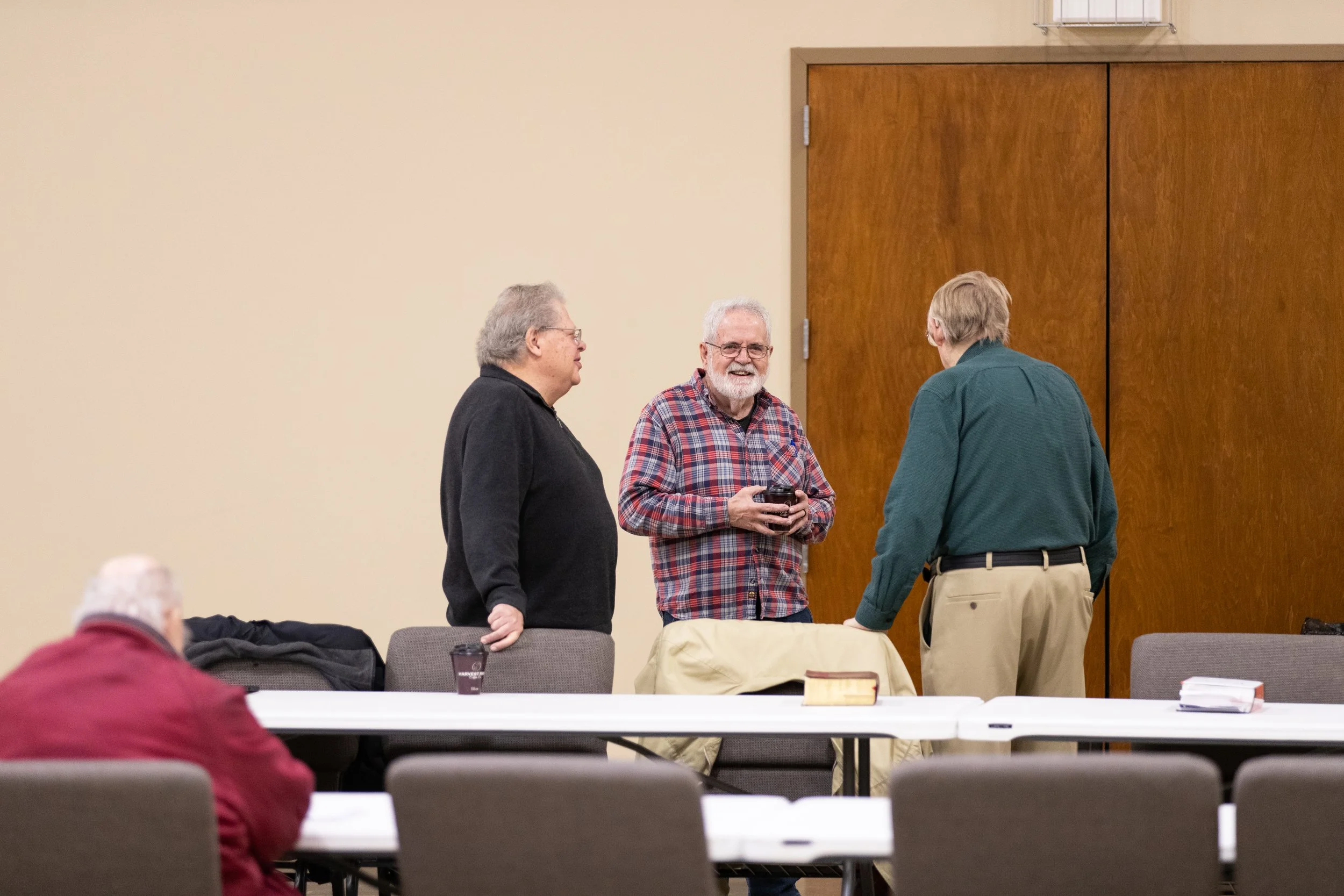 Three elderly men standing and talking in a room with tables and chairs, one man is sitting and facing away from the camera.
