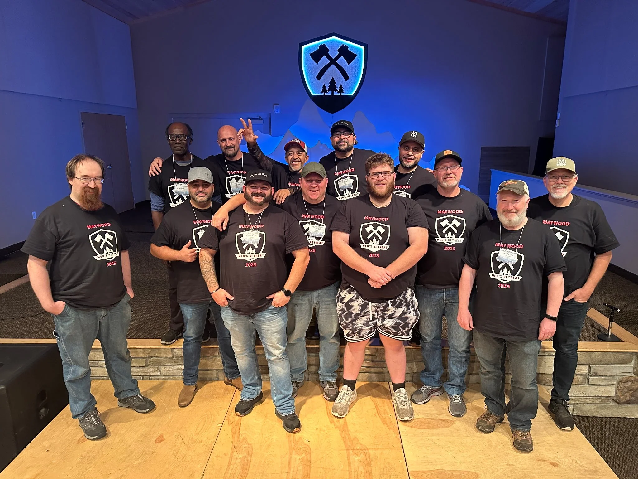 Group of men standing on a stage wearing matching black T-shirts with a white logo and red text, with a shield emblem with crossed axes in the background. They are smiling, some with arms around each other, posing for a photo.