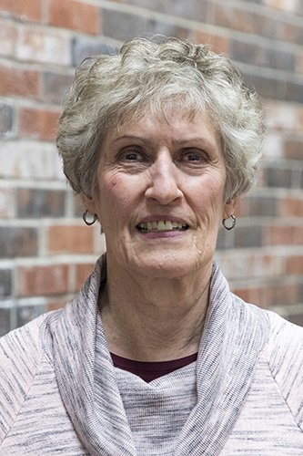 An elderly woman with short, curly gray hair smiling in front of a brick wall, wearing a light-colored jacket and hoop earrings.