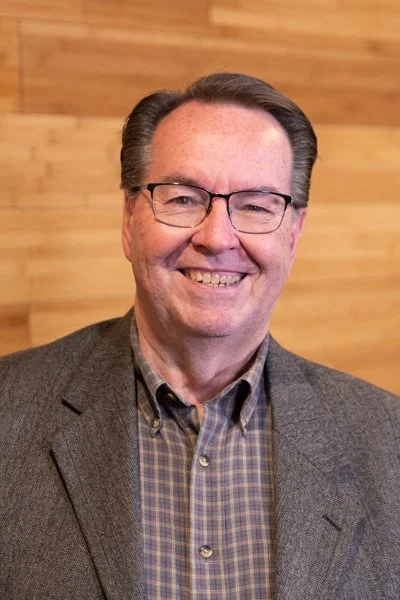 A smiling middle-aged man wearing glasses, a gray blazer, and a plaid shirt, standing in front of a wooden wall.