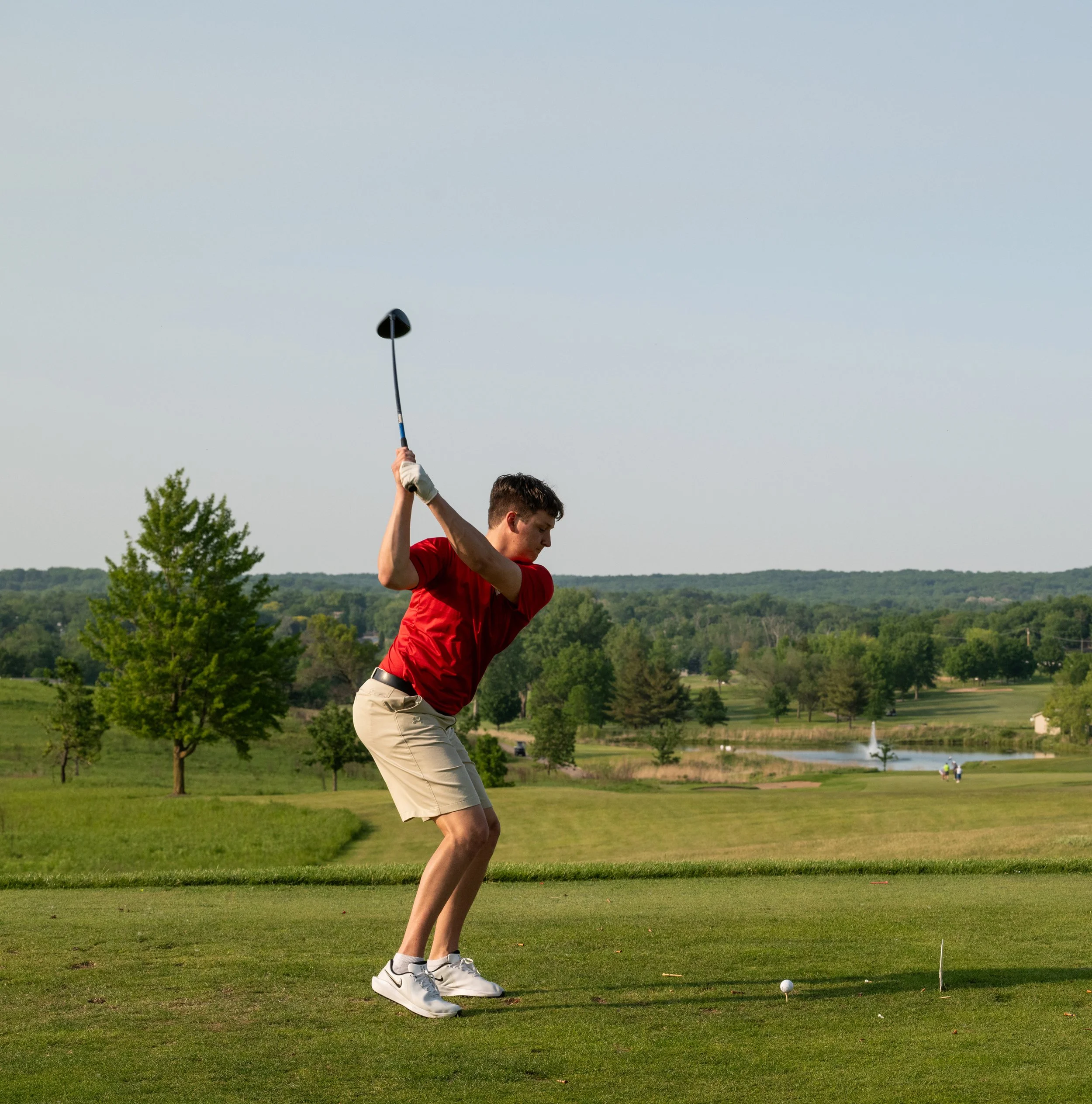 A man in a red shirt and beige shorts preparing to swing a golf club on a golf course with green grass, trees, and a pond in the background.