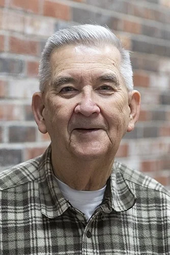 Portrait of an elderly man with gray hair, wearing a plaid shirt, standing outdoors in front of a brick wall.