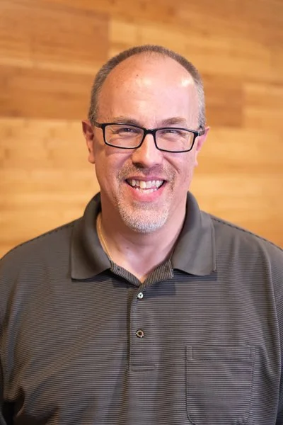 A smiling man with glasses and short hair, wearing a dark gray polo shirt, standing in front of a wooden wall.