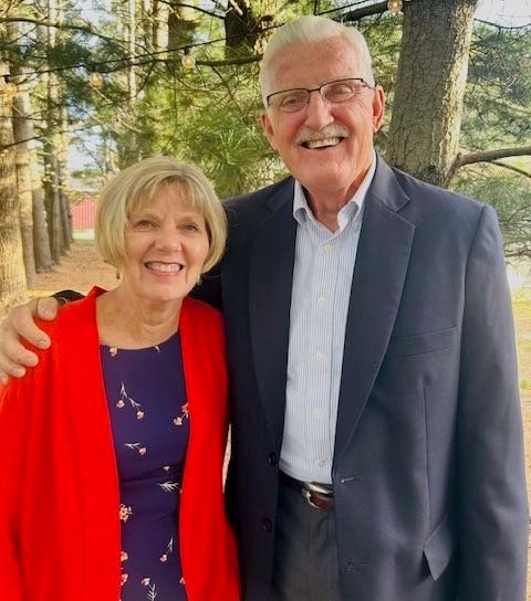 Smiling elderly couple outdoors in a wooded area, with the man wearing glasses and a blue blazer, and the woman in a red jacket and purple dress with a floral pattern.