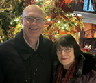 A smiling man and woman standing together indoors near a decorated Christmas tree.