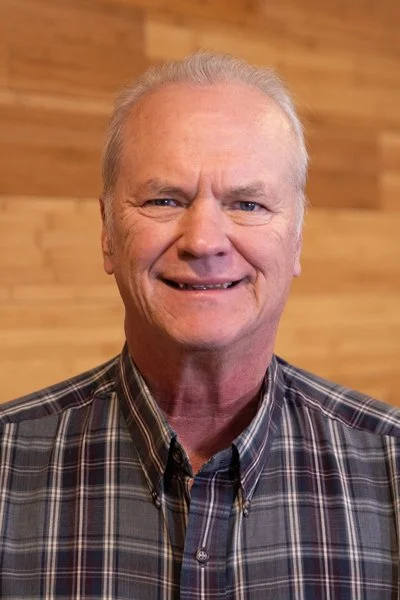 A smiling elderly man with gray hair, wearing a plaid shirt, standing in front of a wooden wall.