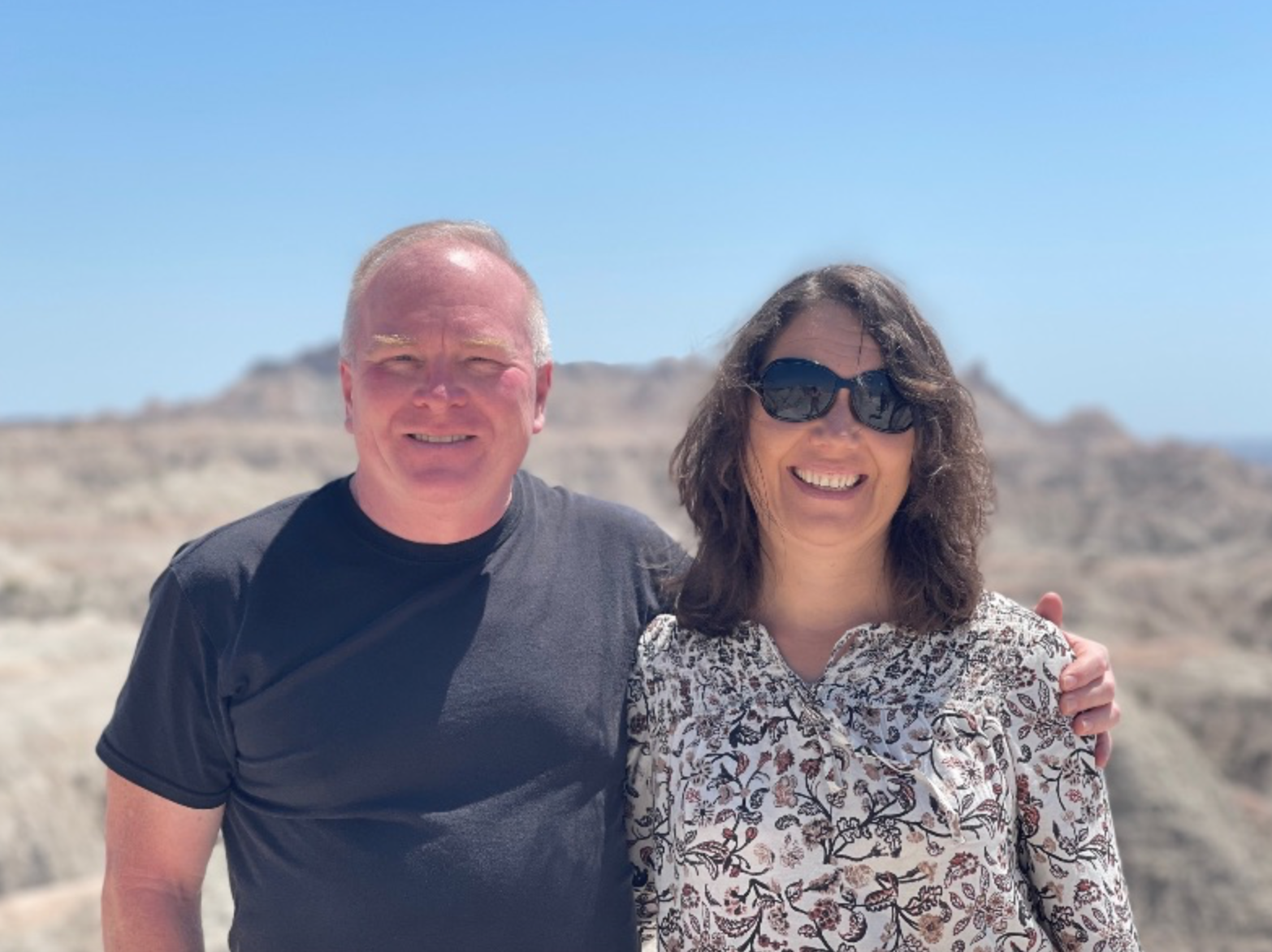 A smiling man and woman standing outdoors on a bright, sunny day with a clear blue sky and rocky landscape in the background. The man has short blond hair and is wearing a black T-shirt. The woman has medium-length brown hair, sunglasses, and is wearing a patterned blouse.