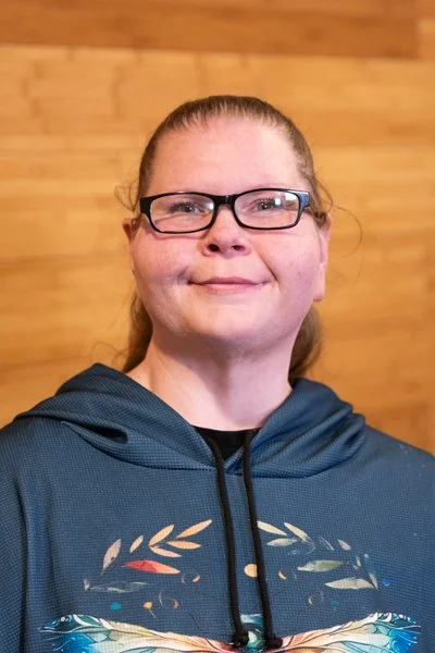 A woman with glasses and red hair tied back, wearing a dark blue hoodie with a butterfly design, standing in front of a wooden wall.