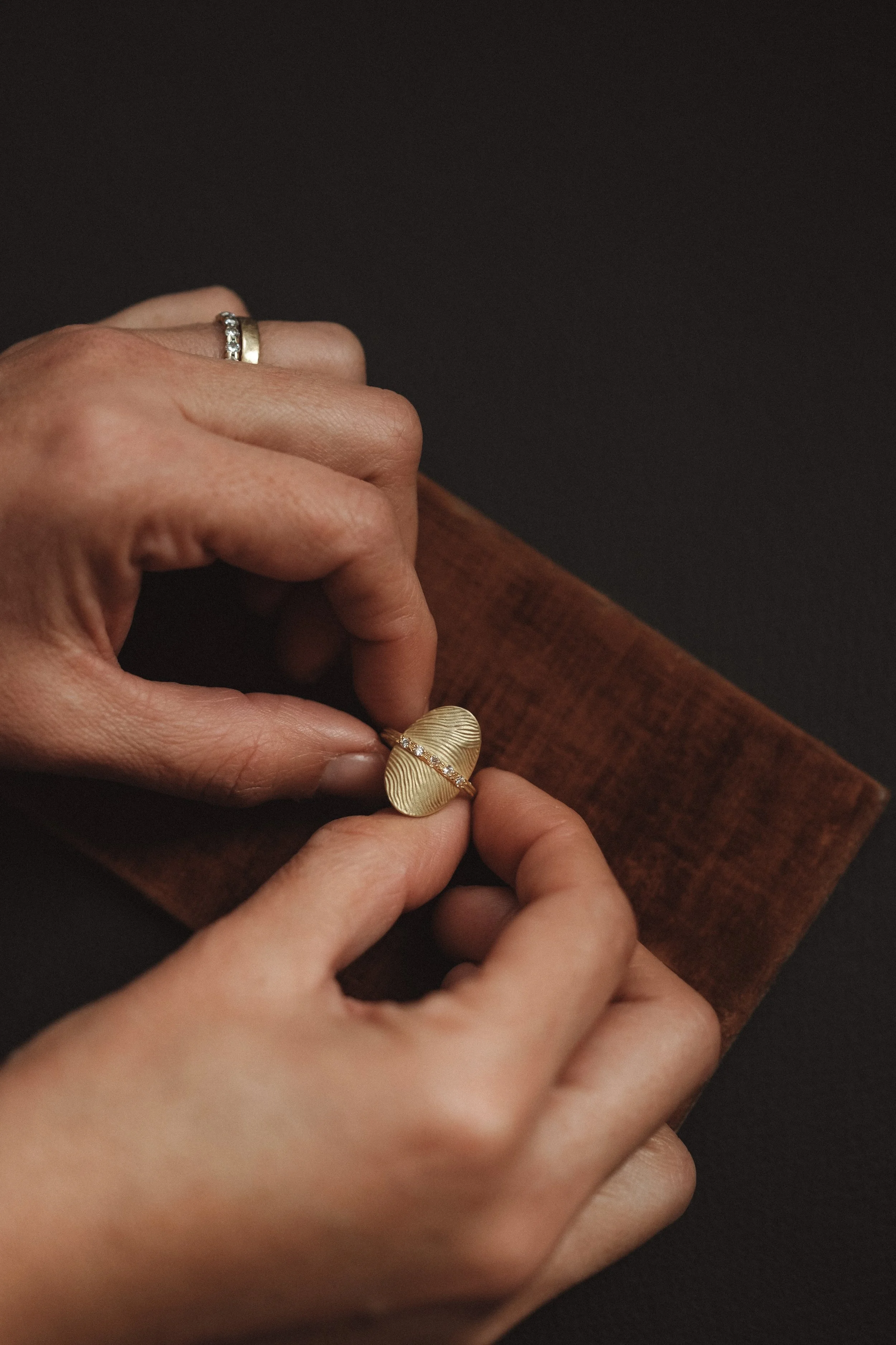 Mains féminines manipulant une bague dorée avec une pierre précieuse, posée sur un support en bois, fond noir.