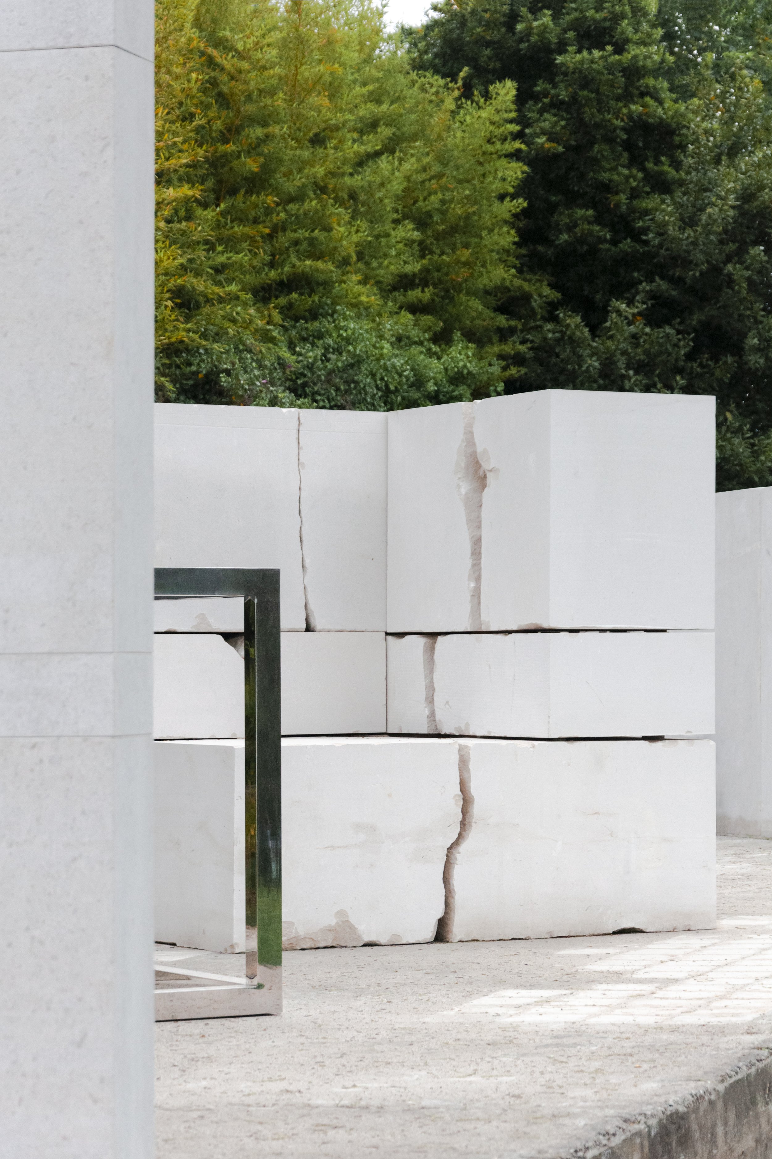 A white stone wall with visible cracks, situated outdoors with green trees in the background, and a metal frame to the left.