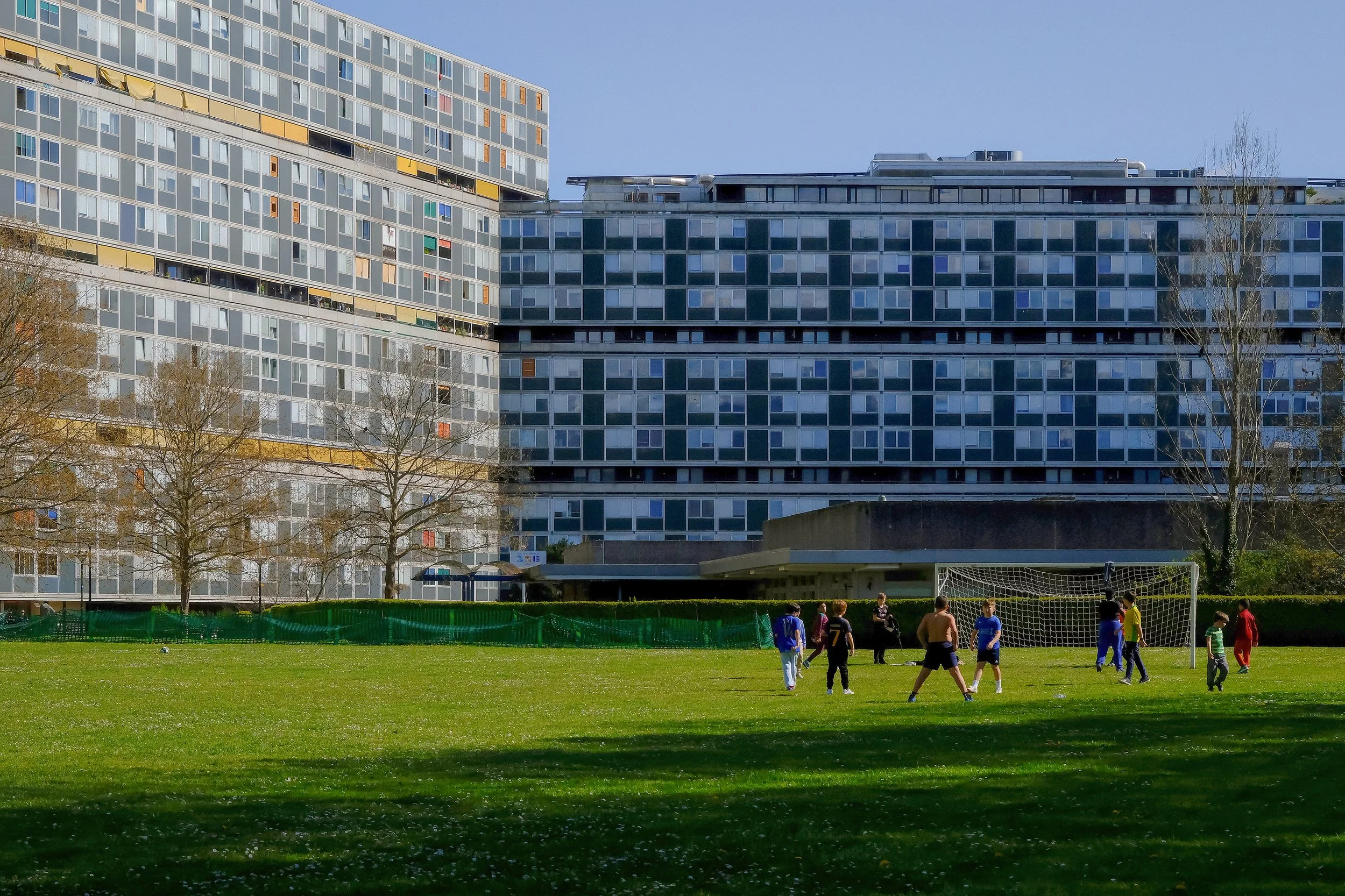 People playing soccer on a grassy field with modern multi-story apartment buildings in the background.