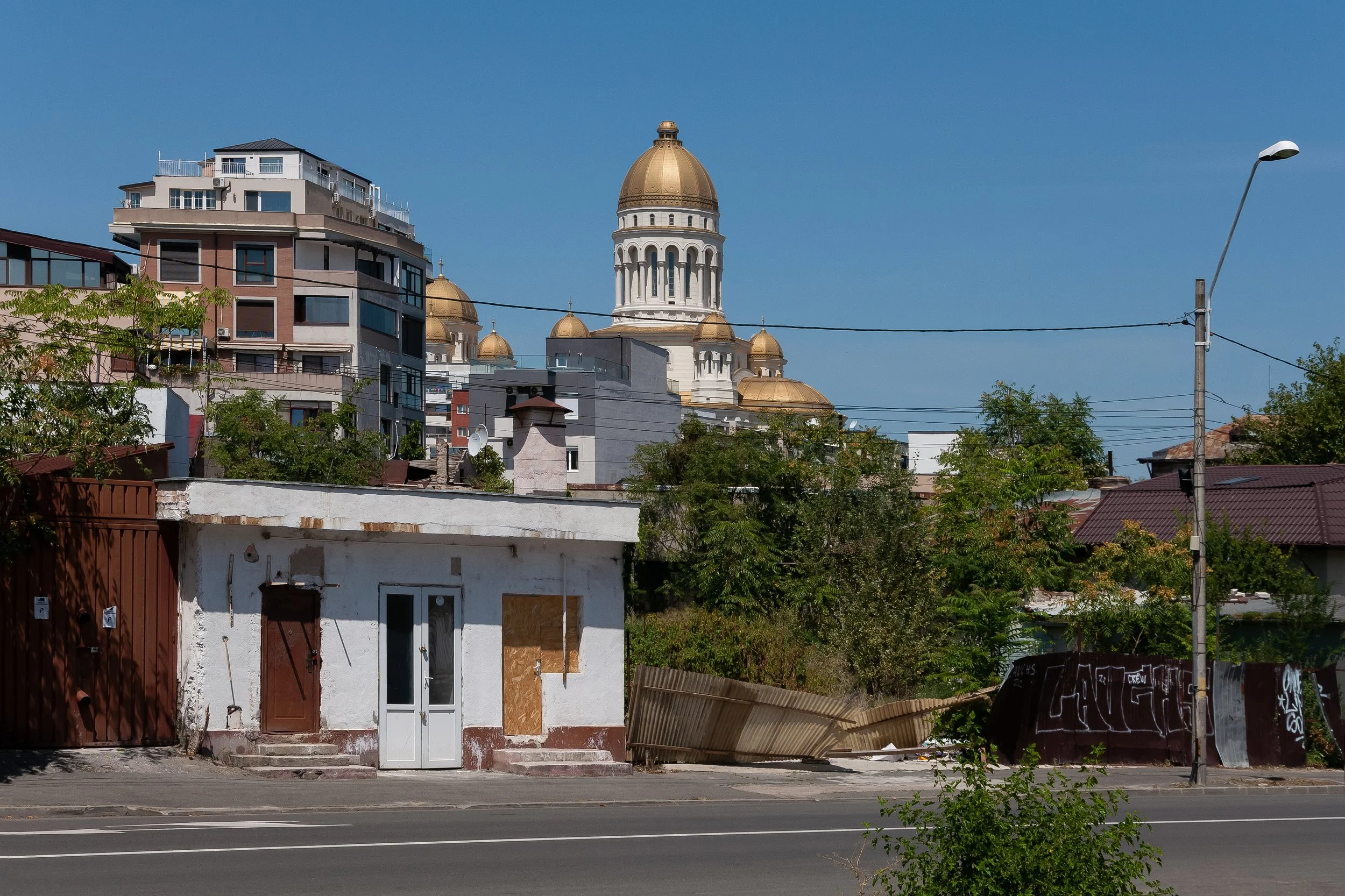 A small weathered white building with a door and boarded-up window in the foreground, a metal fence to the right, and a large church with golden domes and a tall tower in the background, under a clear blue sky.