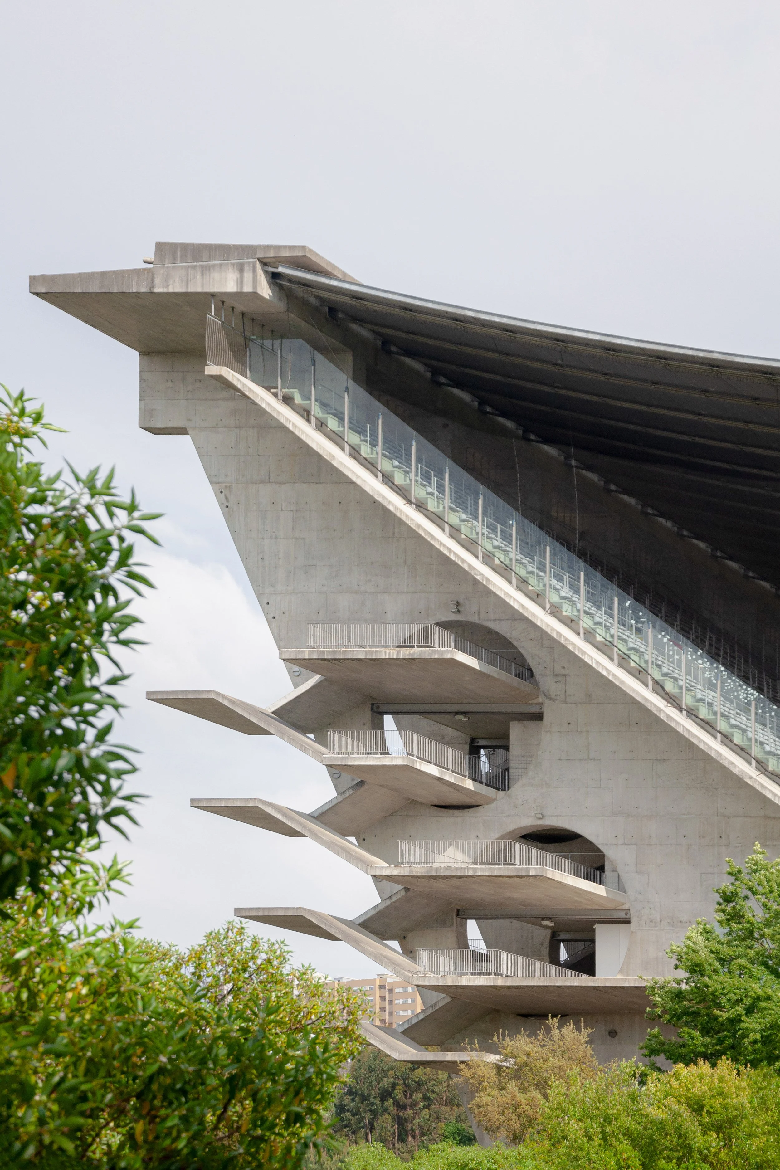 Modern concrete building with tiered, cantilevered floors, surrounded by greenery and a cloudy sky.