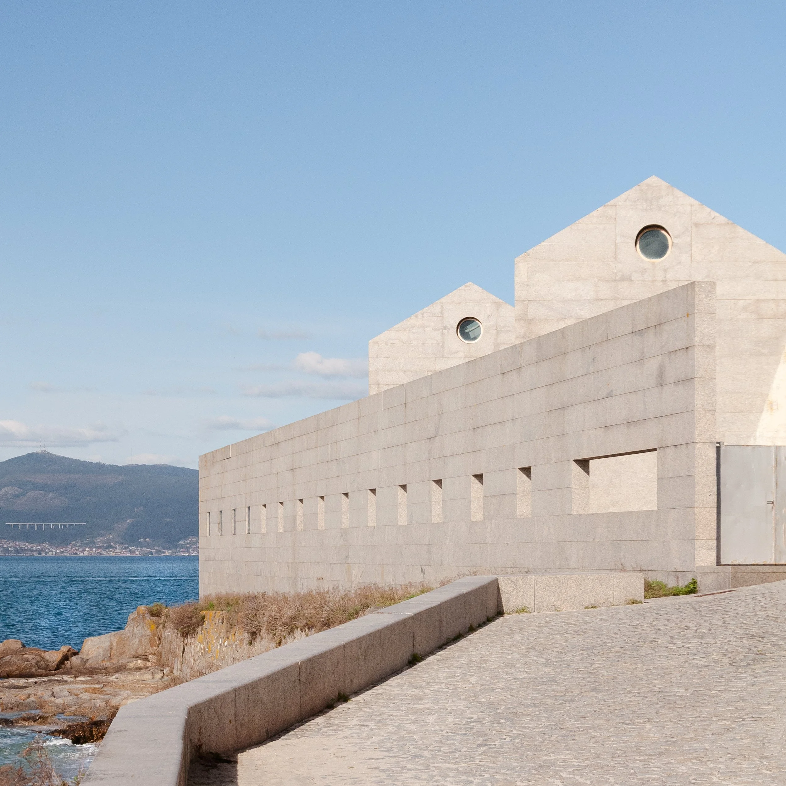 A modern stone building with circular windows and sloped roofs along a coastal walkway, with water and hills in the background.