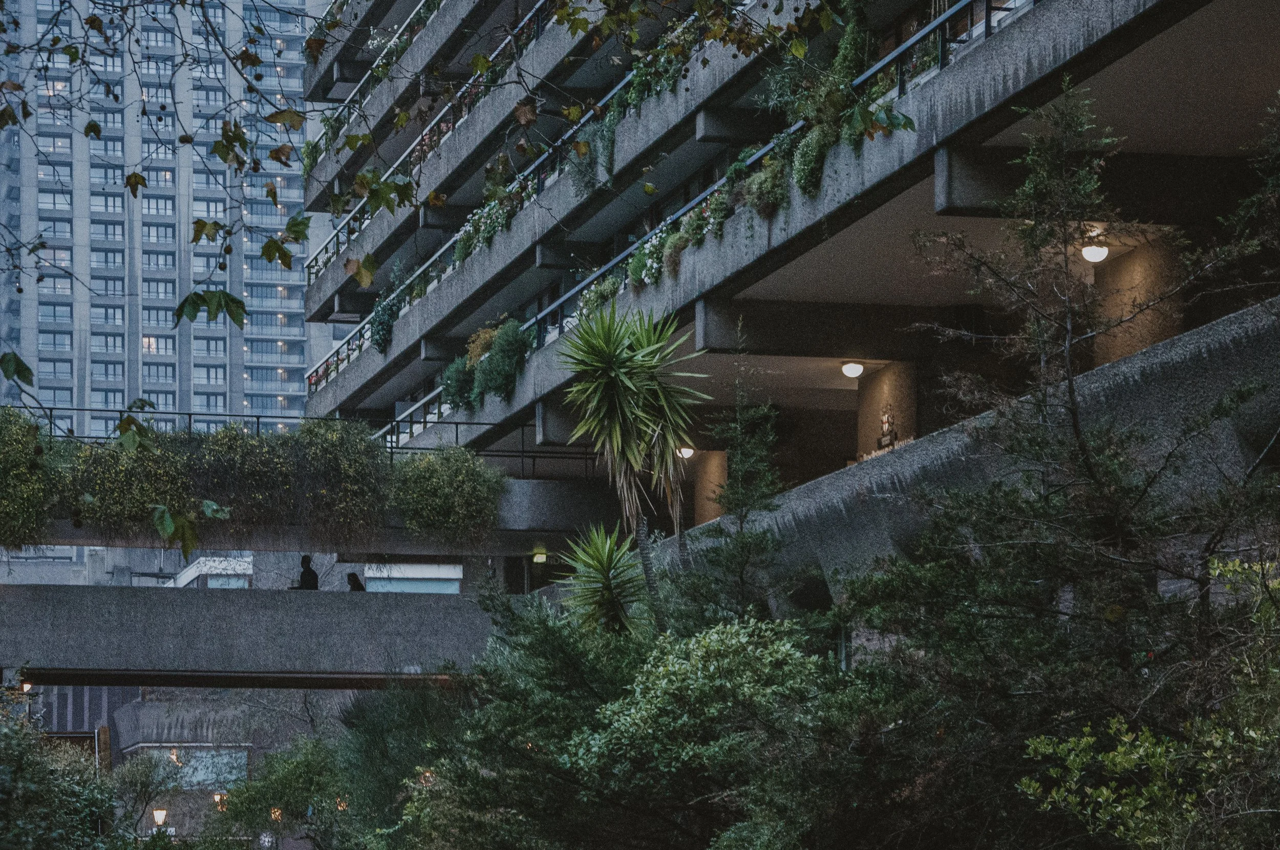 Urban apartment building with balconies decorated with plants, trees, and outdoor lighting, with high-rise buildings in the background.