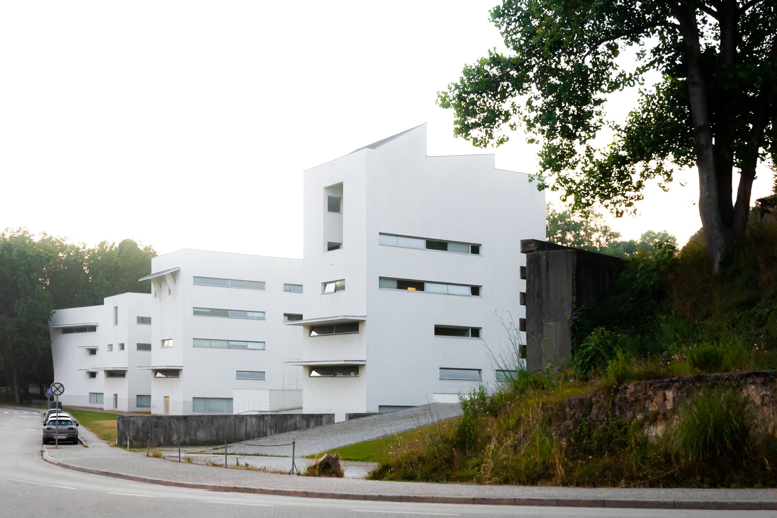 Modern white building with geometric design, narrow horizontal windows, located next to a tree and a grassy area, under a bright sky.
